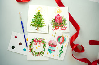 Four painted DIY watercolor Christmas cards: tree, candle, wreath and ornaments, with paint dots, a brush and red ribbon