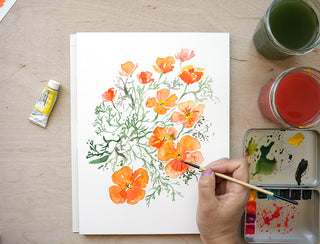 A hand holding a paintbrush is painting california poppies on a white sheet of paper, arranged on a light wood desk with watercolor paints, jars of paint, a tube of paint, and pencils.
